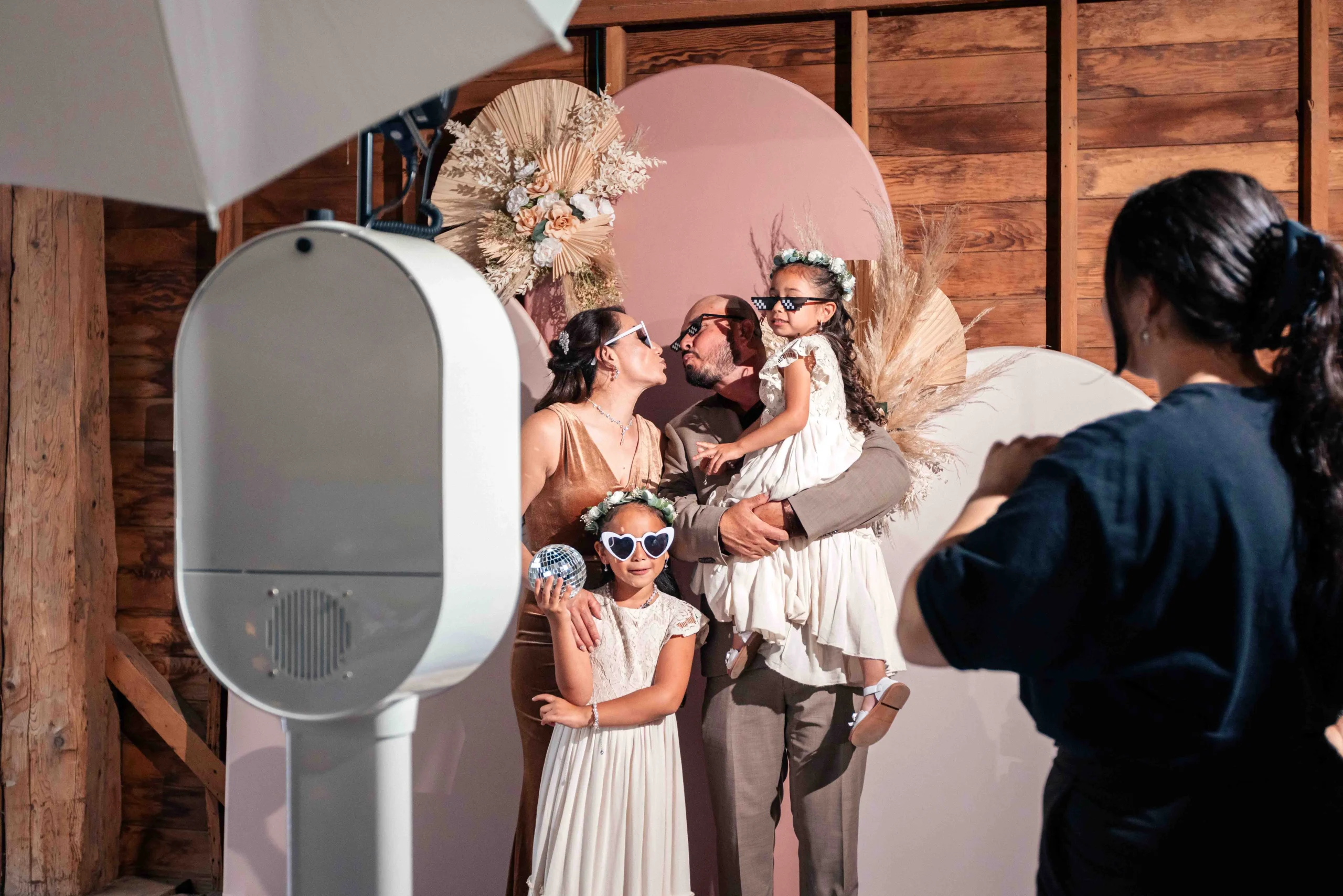 Family posing for a photo booth, with two children in front. A PREVERIE photo booth co. specialist is adjusting the setup in a rustic wooden setting.