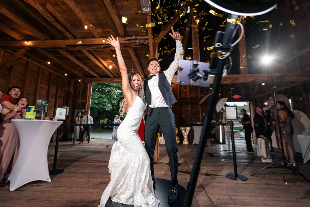 Bride and groom celebrate joyfully with arms raised in front of a photo booth station