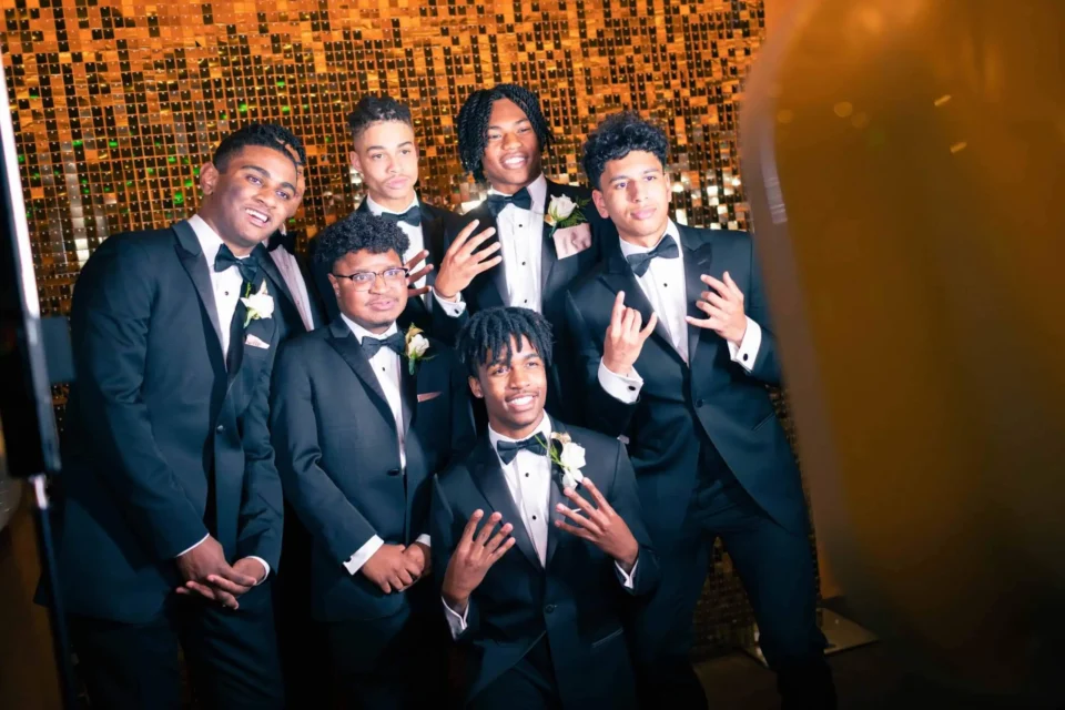 Six young men in tuxedos pose together in front of a golden sequin backdrop and smiling at the photo booth camera.