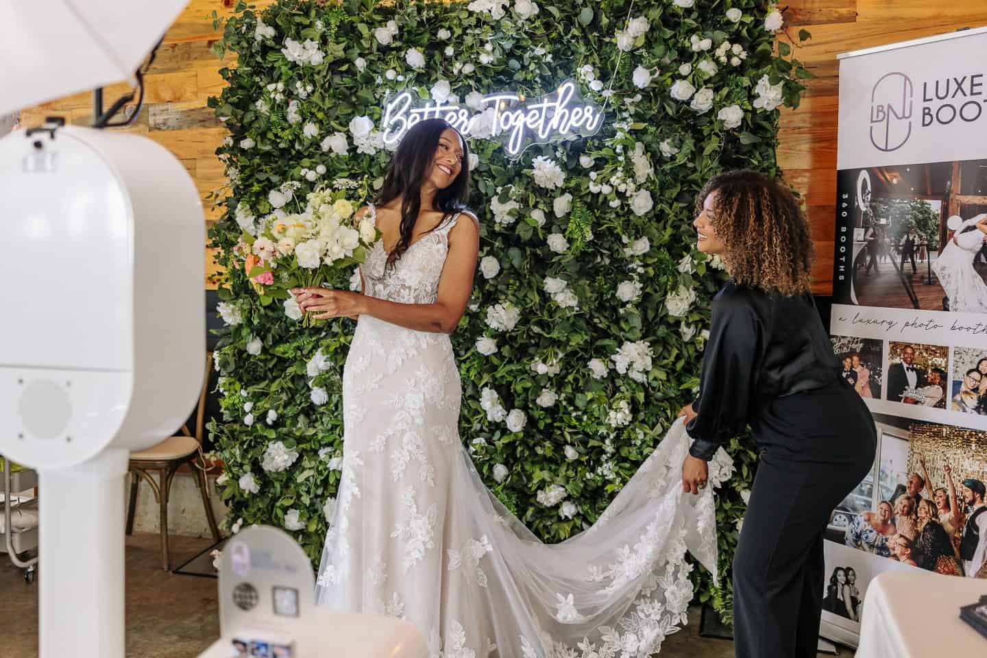 A bride in a lace gown stands smiling in fornt of a professional photo boot station with a floral backdrop behind her