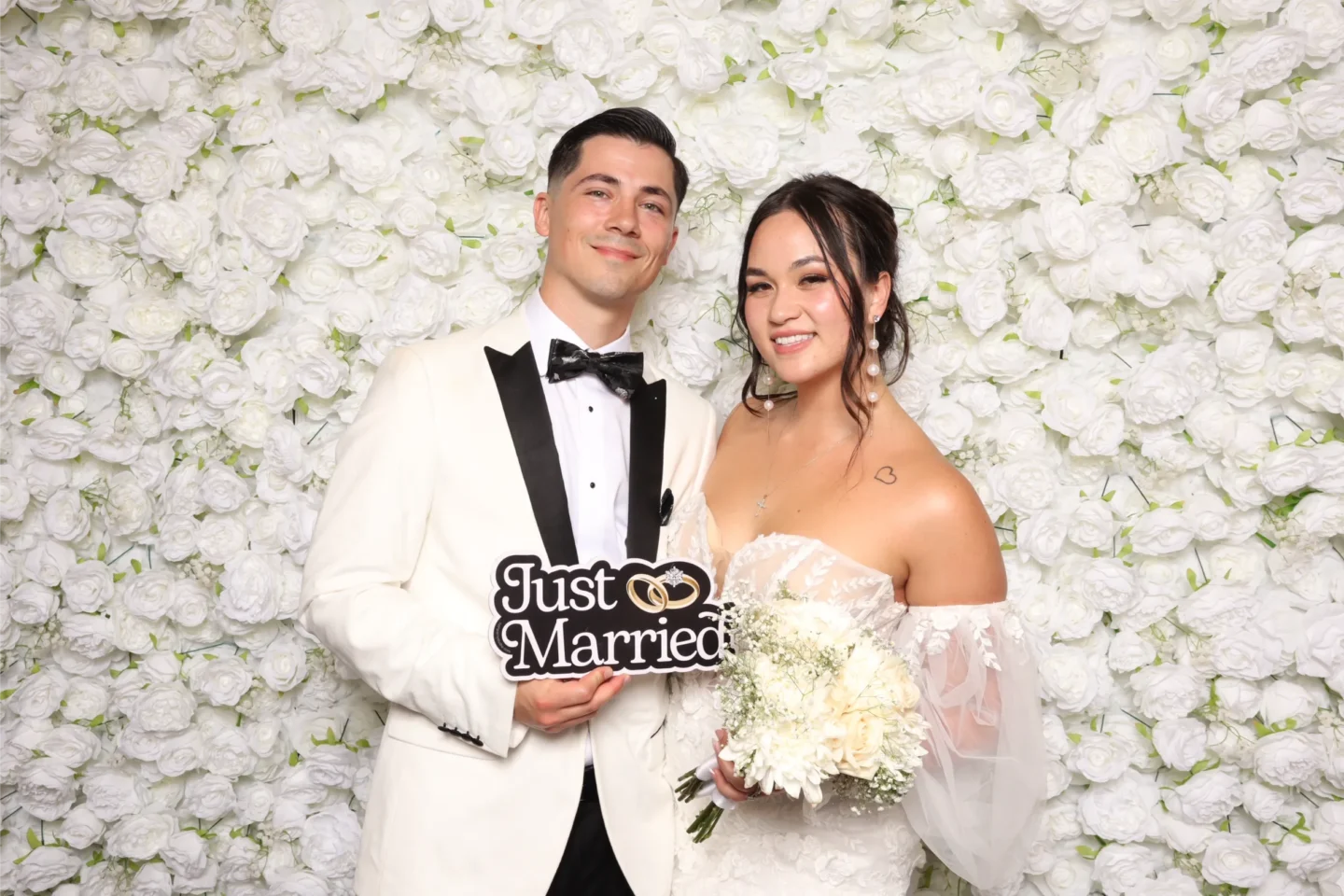 A bride and groom pose in front of a wall of white roses.