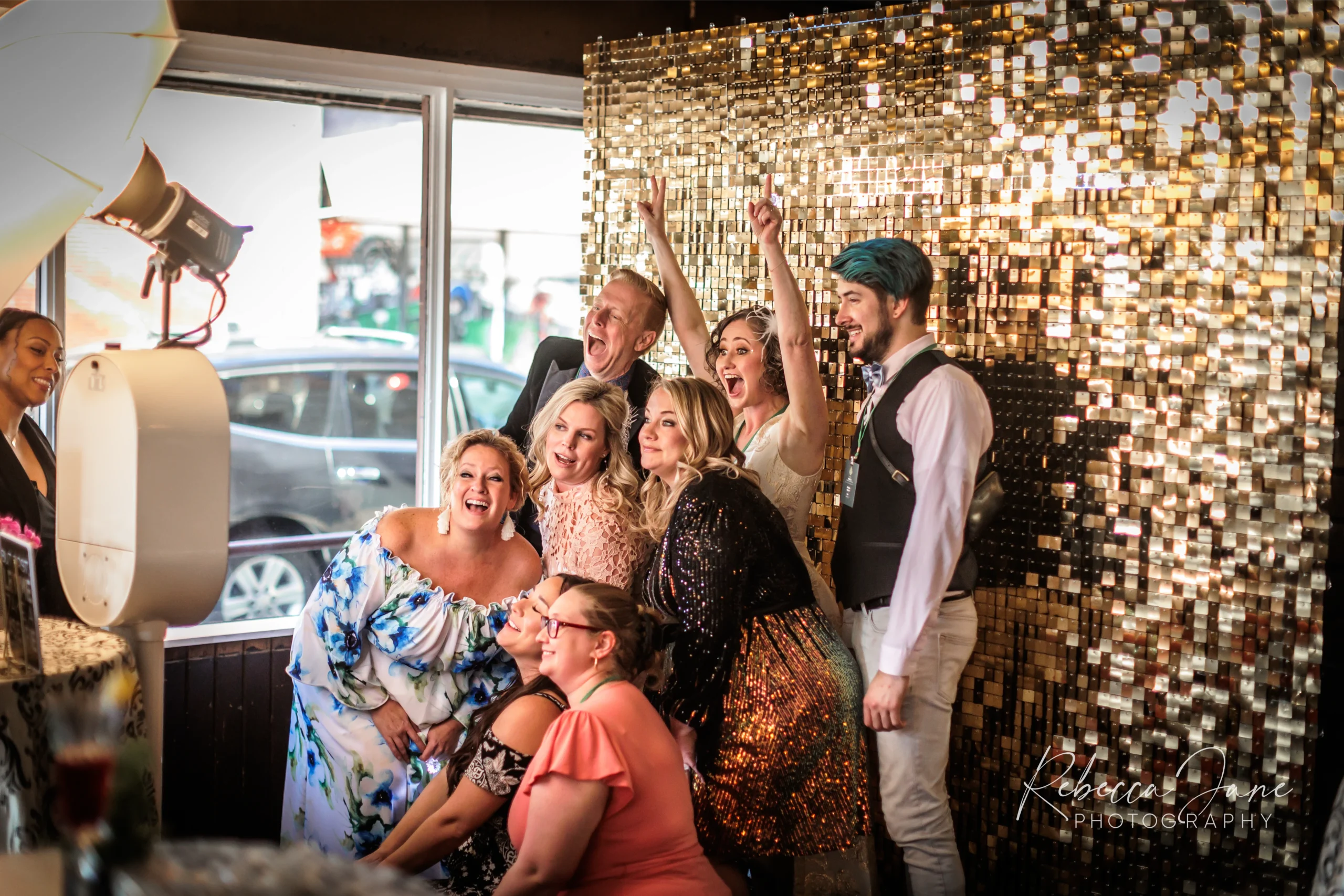 A group of people posing joyfully in front of a gold sequin backdrop at an indoor event.