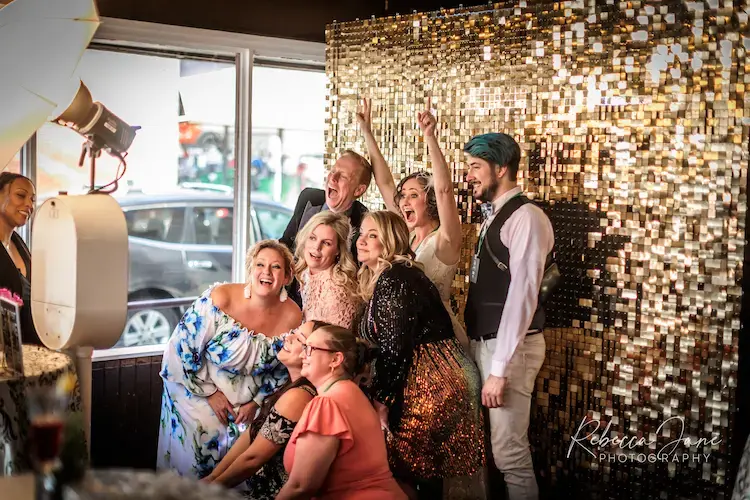 A group of people posing joyfully in front of a gold sequin backdrop at an indoor event.