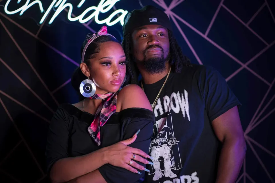 A woman and a man pose together in front of a geometric-patterned wall with a neon sign.