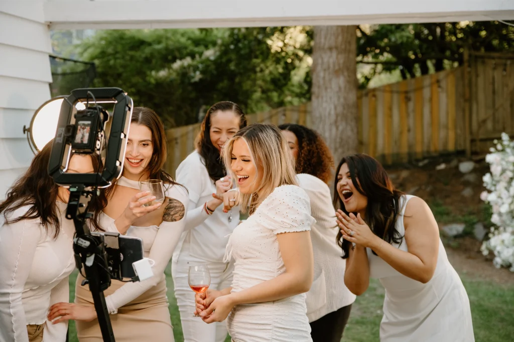 A group of women in white outfits stand outdoors and looking at a photo booth camera on a tripod