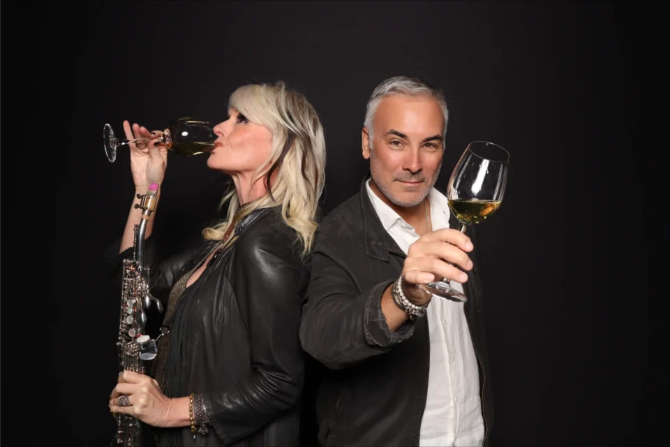 A woman holding a saxophone drinks wine, while a man holding a glass of wine raises it in front of him.