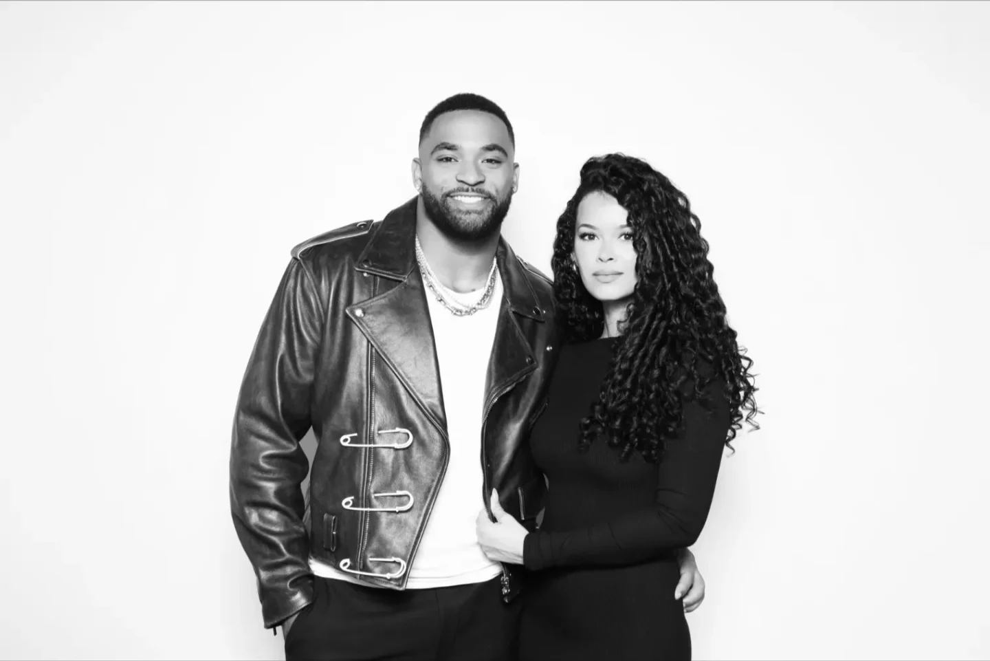 A man in a leather jacket and a woman in a black dress pose together against a plain background.