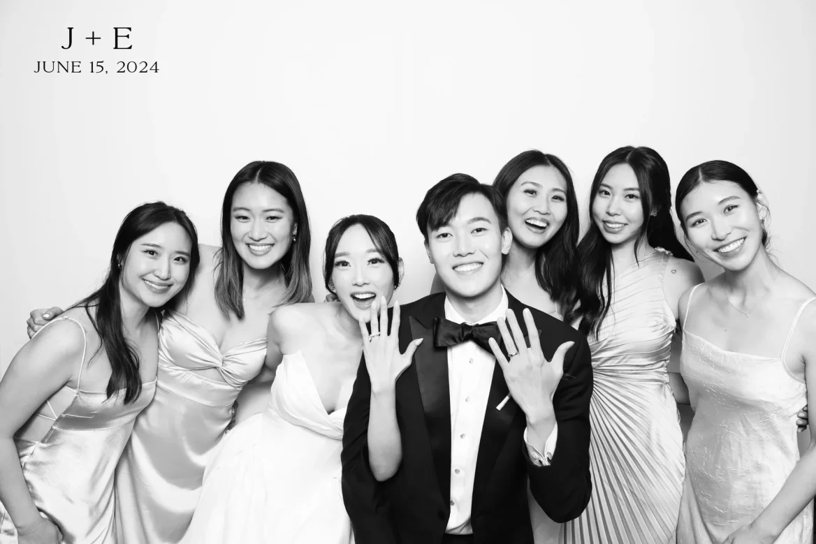 A group of seven people in formal attire pose together in front of a photo booth camera.