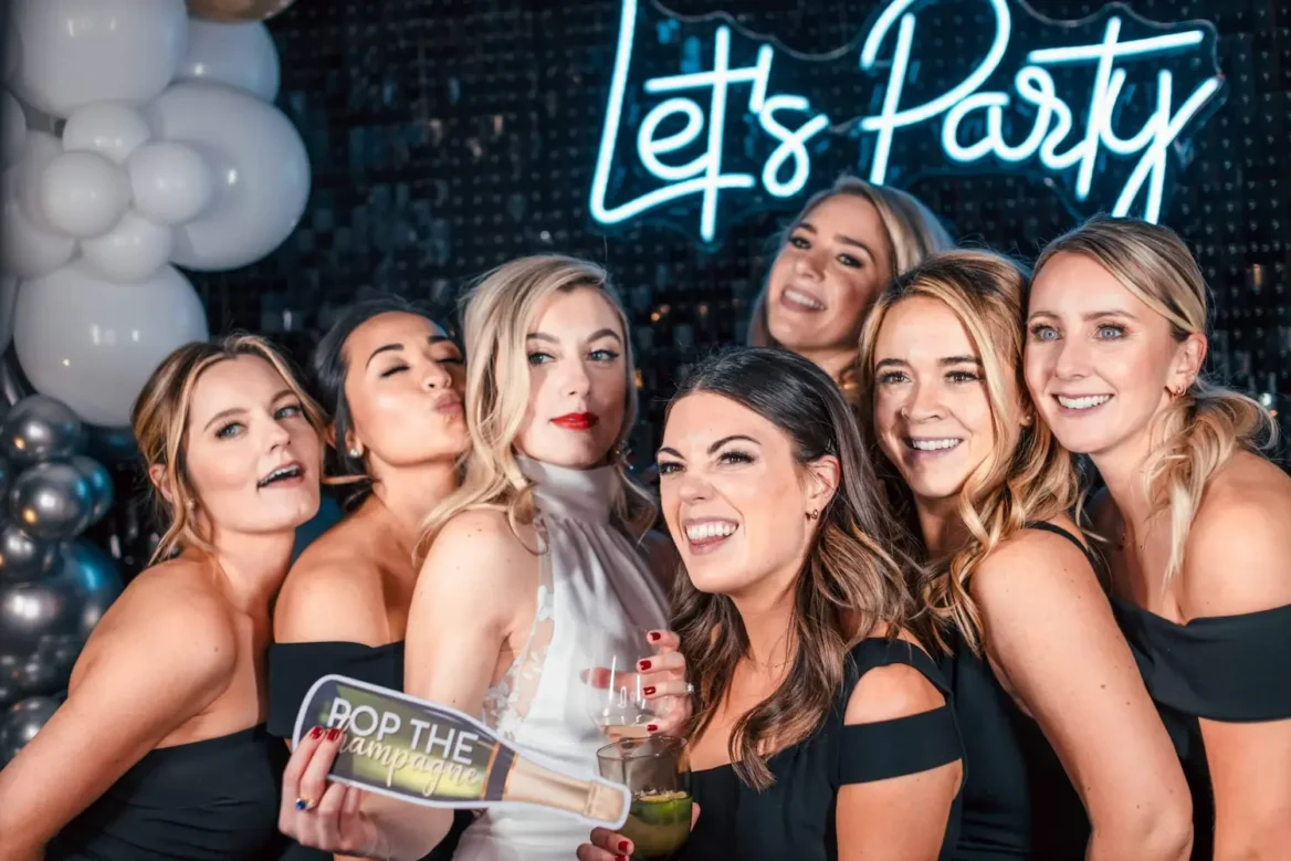 A group of women smiling and laughing, surrounding the bride in front of a photo booth camera.