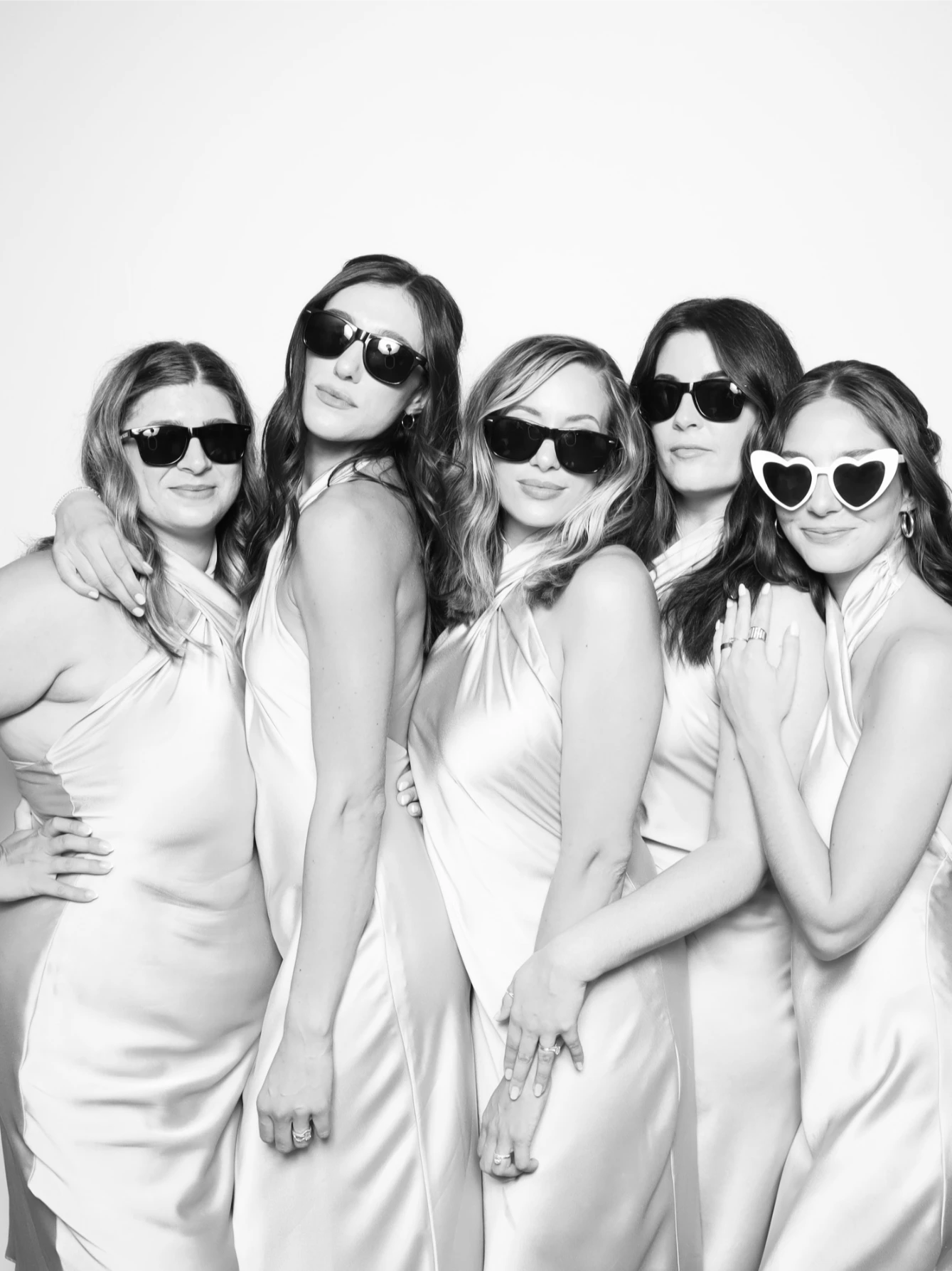 Five women in matching satin dresses and sunglasses pose together against a plain backdrop.